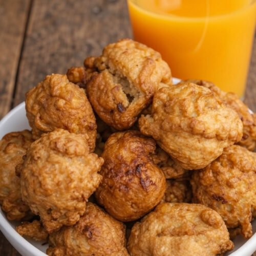 Bol de beignets à la banane moelleux et dorés, fraîchement frits, posé sur une table en bois rustique. En arrière-plan, un verre de jus de fruits frais et tropical, suggérant un jus de mangue ou d'orange, illustrant parfaitement la recette de beignets à la banane africains pour un goûter anti-gaspi.